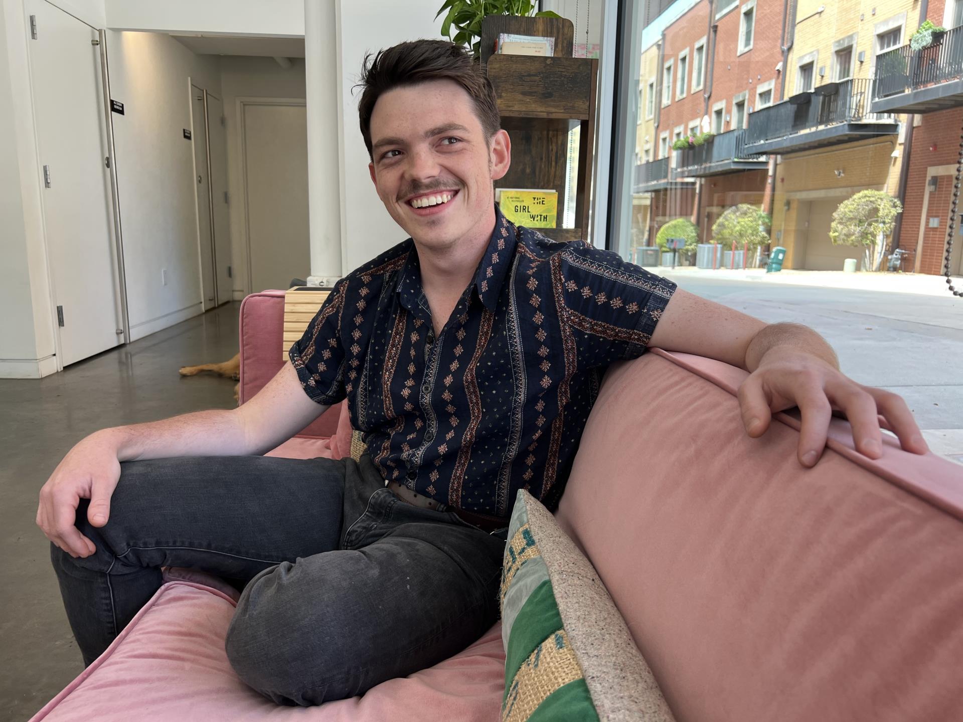 white man with mustache smiling while sitting on pink couch